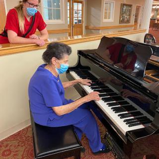 An elderly woman plays the piano while another person watches, both wearing masks in a care facility.