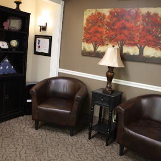 A cozy waiting area featuring two brown leather chairs, a decorative lamp, and artwork of trees on the wall.
