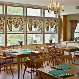 A cozy dining room with wooden furniture and natural light streaming through large windows, adorned with floral curtains.