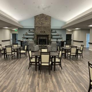 A spacious dining area with wooden flooring, featuring multiple tables and chairs, centered around a fireplace with surrounding shelves.