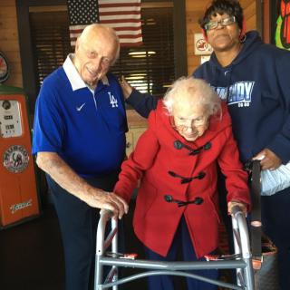 Three individuals pose together indoors, with an elderly woman using a walker.