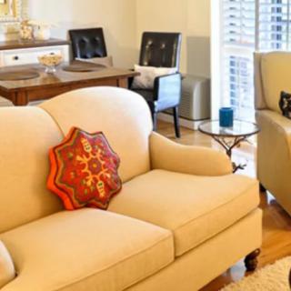 A cozy living room featuring a beige sofa with a decorative pillow, accompanied by a wooden table and bright window light.