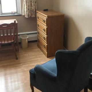 A simple room with a wooden desk, a blue armchair, and a dresser next to a window.
