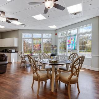 A modern dining area with ample natural light, featuring elegant tables and chairs, large windows, a kitchen counter, and ceiling fans.