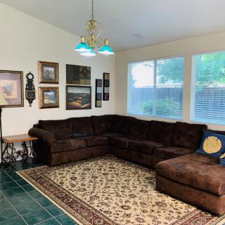 A cozy living room featuring a large brown sectional sofa, decorative rug, and several pieces of wall art. The room is well-lit by natural light coming through the windows.