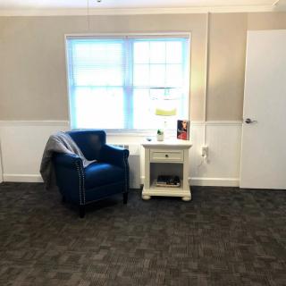 A cozy living room corner with a blue armchair next to a side table and lamp, illuminated by natural light through the window.