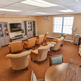 A cozy living room with multiple chairs arranged around a television, featuring carpeted flooring and large windows.
