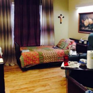 A cozy bedroom features a single bed with a colorful quilt, a side table adorned with personal items, and decor including a cross on the wall and a painting. The room is completed with elegant curtains and wooden flooring.