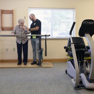 A therapist assists an elderly woman with exercises using parallel bars in a rehabilitation facility.