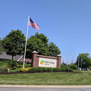 Entrance sign for a community or facility with an American flag and landscaped surroundings.