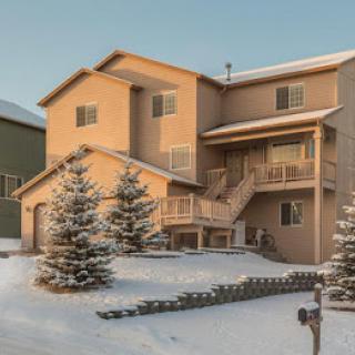 A snow-covered house in a residential area, illuminated by soft sunlight, with evergreen trees in the front yard.