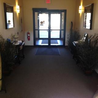 A well-lit hallway leading to a glass door entrance, adorned with plants and decorated with framed artwork on the walls.