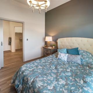 A cozy bedroom featuring a floral-patterned bed with decorative pillows, a nightstand with a lamp, and a stylish chandelier.