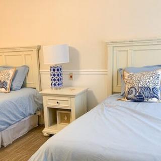 A cozy bedroom with two twin beds featuring blue bedding and decorative pillows, separated by a white nightstand with a lamp.