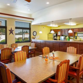 A cozy dining area with wooden chairs and tables, adjacent to a kitchen, featuring warm lighting and large windows.