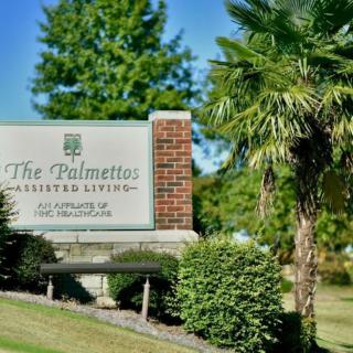 A sign for The Palmettos Assisted Living surrounded by lush greenery and palm trees.