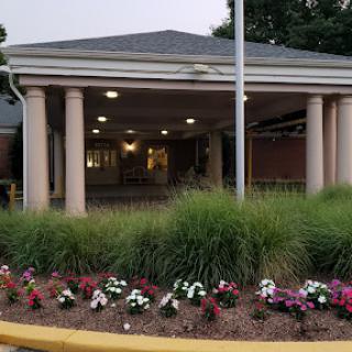 The entrance of a building surrounded by well-maintained flower beds and greenery, with columns and a flagpole.