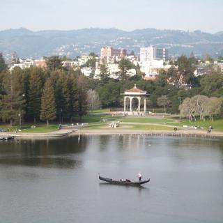 A scenic view of a lake with a boat, surrounded by a park and city buildings in the background.
