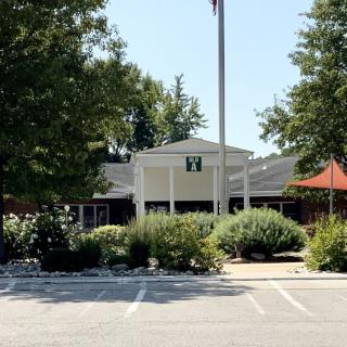 The image shows the exterior of a building surrounded by trees and landscaping, with a parking lot in front.
