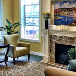 A cozy living room featuring an armchair and a fireplace, adorned with a painting and surrounded by plants.