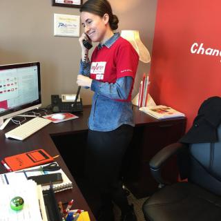 A woman is on the phone in an office setting, standing by a desk with a computer.