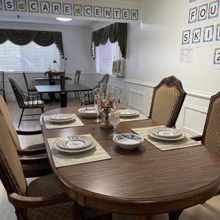 A dining room with a neatly set table, featuring plates and cutlery, surrounded by cushioned chairs and flanked by windows with curtains.