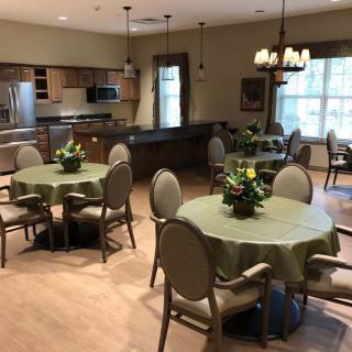 A cozy dining area with tables and chairs, featuring a kitchen in the background and decorative plants on the tables.