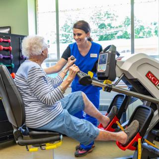A healthcare professional assists an elderly woman with exercise using specialized equipment in a physical therapy setting.
