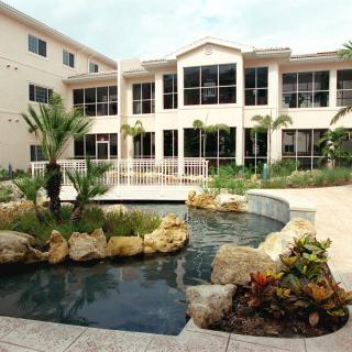 A residential building with a landscaped garden and pond in the foreground, featuring palm trees and rocks.