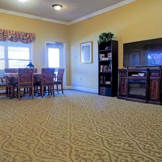 A spacious living room with a dining area, featuring a patterned carpet, television, and bookshelf.