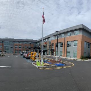 A modern hospital building featuring a flagpole in the parking area, set against a cloudy sky.
