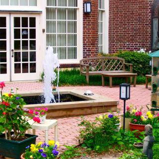 A charming garden patio with a fountain, flowers, and a bench near a brick building.