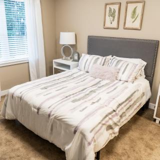 A cozy bedroom with a neatly made bed, featuring striped bedding and a gray headboard. The room includes nightstands with lamps and framed artwork above the bed.