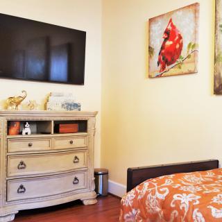 A cozy bedroom featuring a television mounted above a dresser, with colorful artwork and vibrant orange bedding.