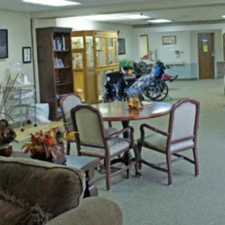 A common area in a nursing home with a table, chairs, and a person in a wheelchair.