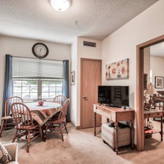 Cozy dining area with a table set for four, featuring a wall-mounted clock and a television in an adjoining living space.