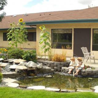 A serene backyard with a pond, sunflowers, and patio seating against the backdrop of a house.