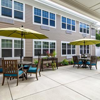 A patio area with tables and chairs, featuring large umbrellas, outside a building with multiple windows.