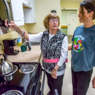 An elderly woman receives help from a caregiver in the kitchen.