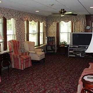 A cozy living room with patterned wallpaper and carpet, featuring multiple chairs, a television, and large windows.