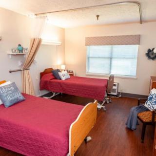 A cozy nursing home bedroom with two beds featuring red bedding and a window providing natural light.