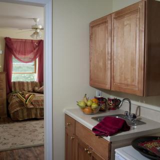View of a kitchen area with wooden cabinets and a sink, leading to a living room with red curtains.