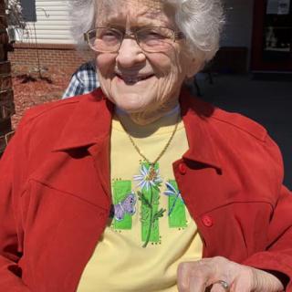 An elderly woman is smiling while sitting outdoors, wearing a red jacket and a yellow shirt.