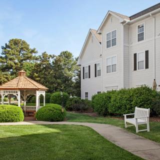 A serene garden area with a gazebo, pathway, and white bench next to a large building.