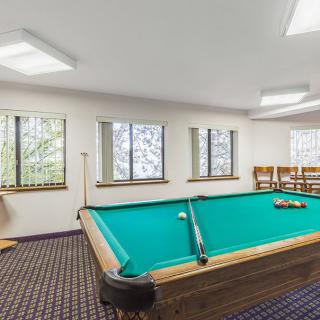 A game room featuring a pool table with cues and balls, surrounded by windows and chairs.