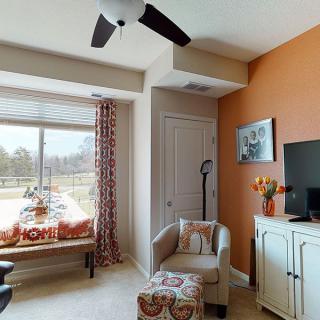 A cozy living room featuring a window seat, armchair, and television, with an orange accent wall and ceiling fan.
