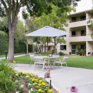 A serene outdoor patio area with tables and chairs under an umbrella, surrounded by flowers and trees, in front of a multi-story building.