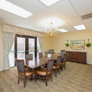 A well-lit dining room with a large conference table and eight chairs, featuring a chandelier, curtains, artwork on the wall, and a sideboard.
