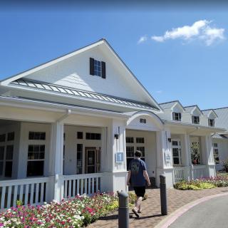 A person walking towards a white building with a porch on a sunny day.