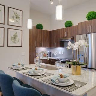 A modern kitchen with a bar counter set for dining, featuring wooden cabinets, stainless steel appliances, and elegant pendant lighting.